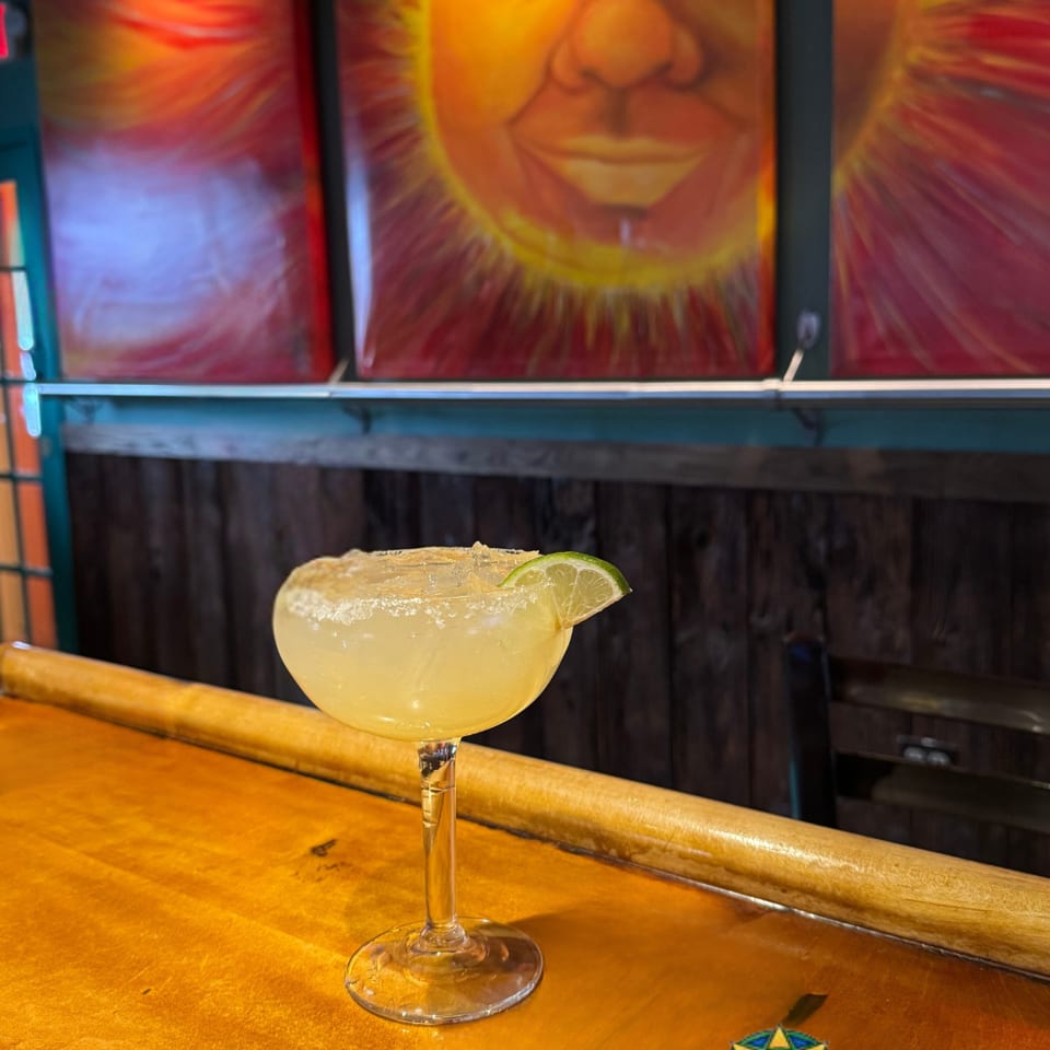 Salt-rimmed margarita with a lime wedge on a wooden bar beneath colorful Tex-Mex artwork.