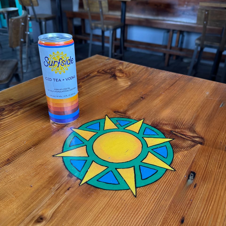 Surfside iced tea and vodka can beside a colorful sun design on a wooden restaurant table.