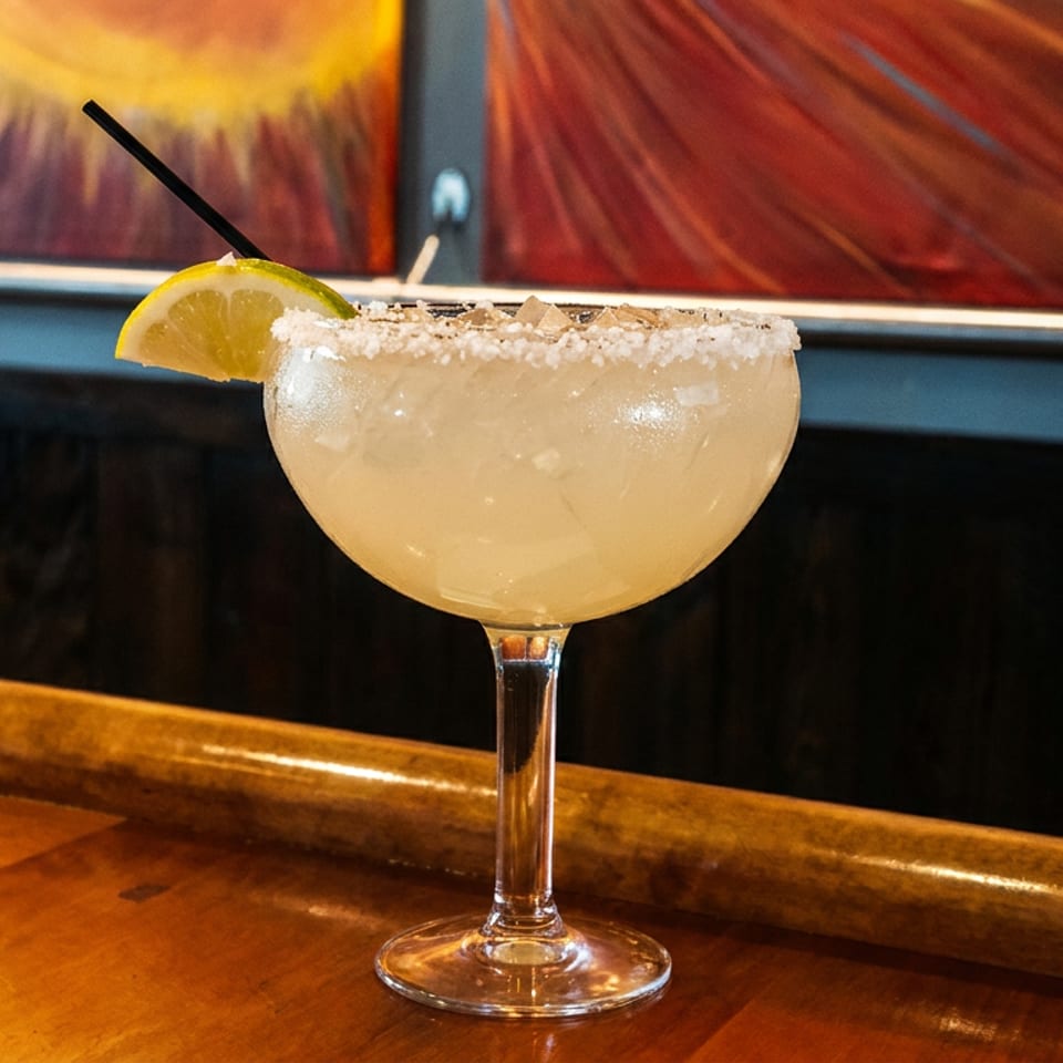 Salt-rimmed margarita garnished with a lime wedge and straw on a polished wooden restaurant bar.