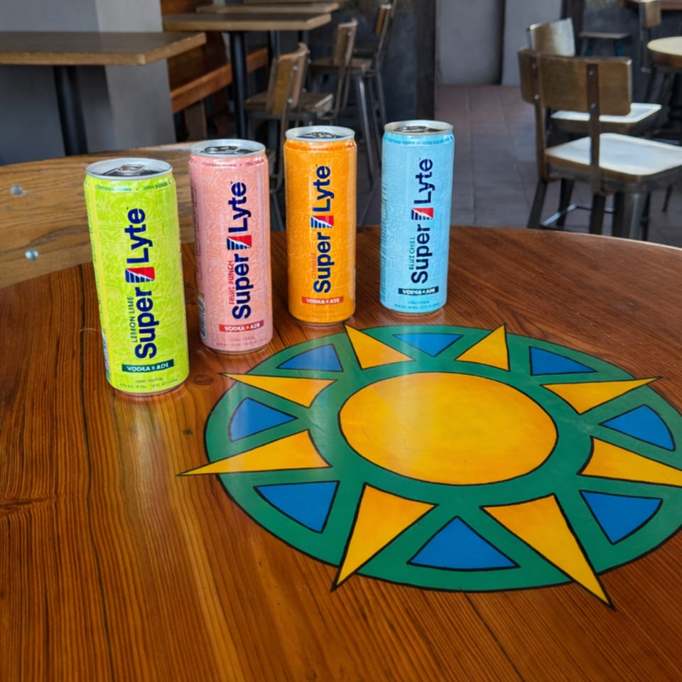 Four colorful Super Lyte canned drinks sit on a wooden restaurant table beside a vibrant geometric sunburst placemat.