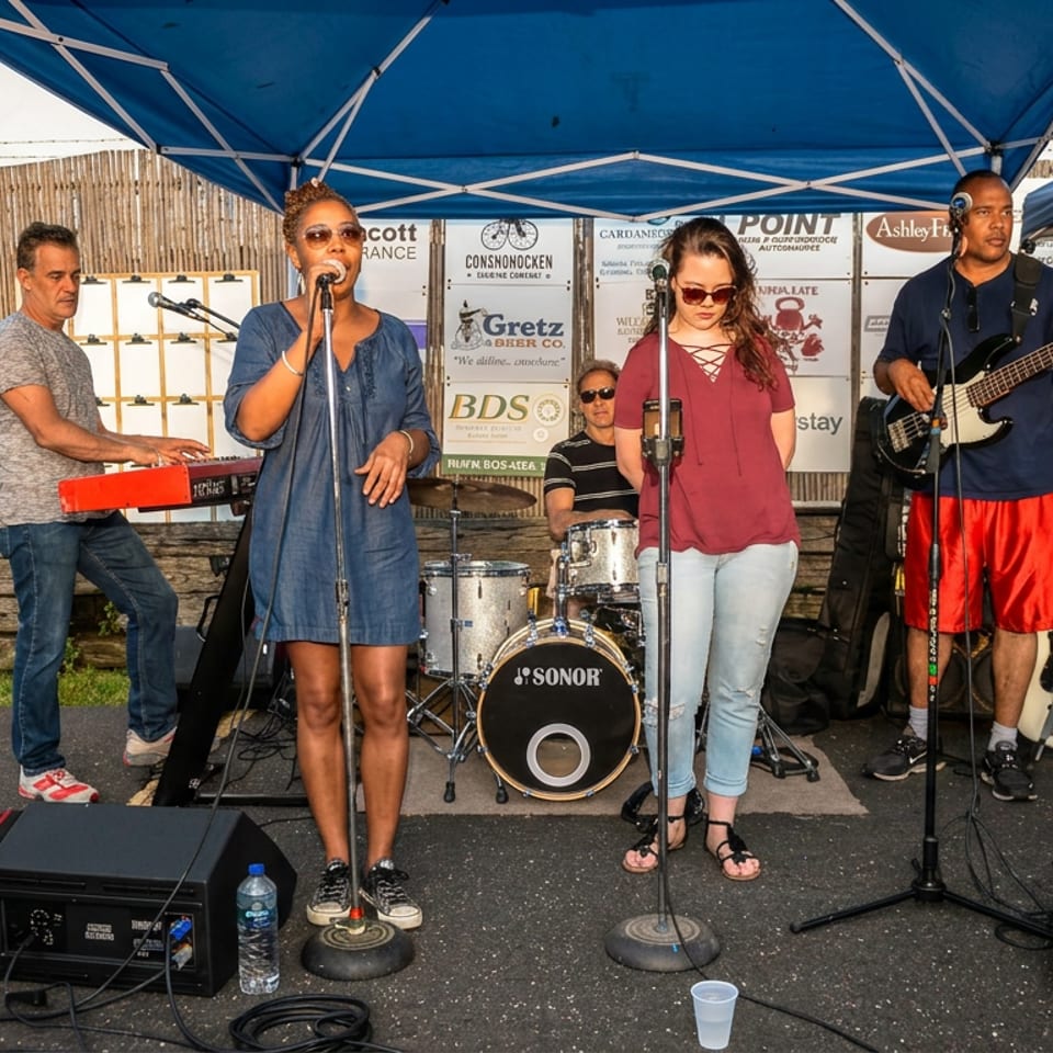 Five-member band performs outdoors under a blue canopy, with singers, keyboards, drums, bass guitar, and sound equipment.
