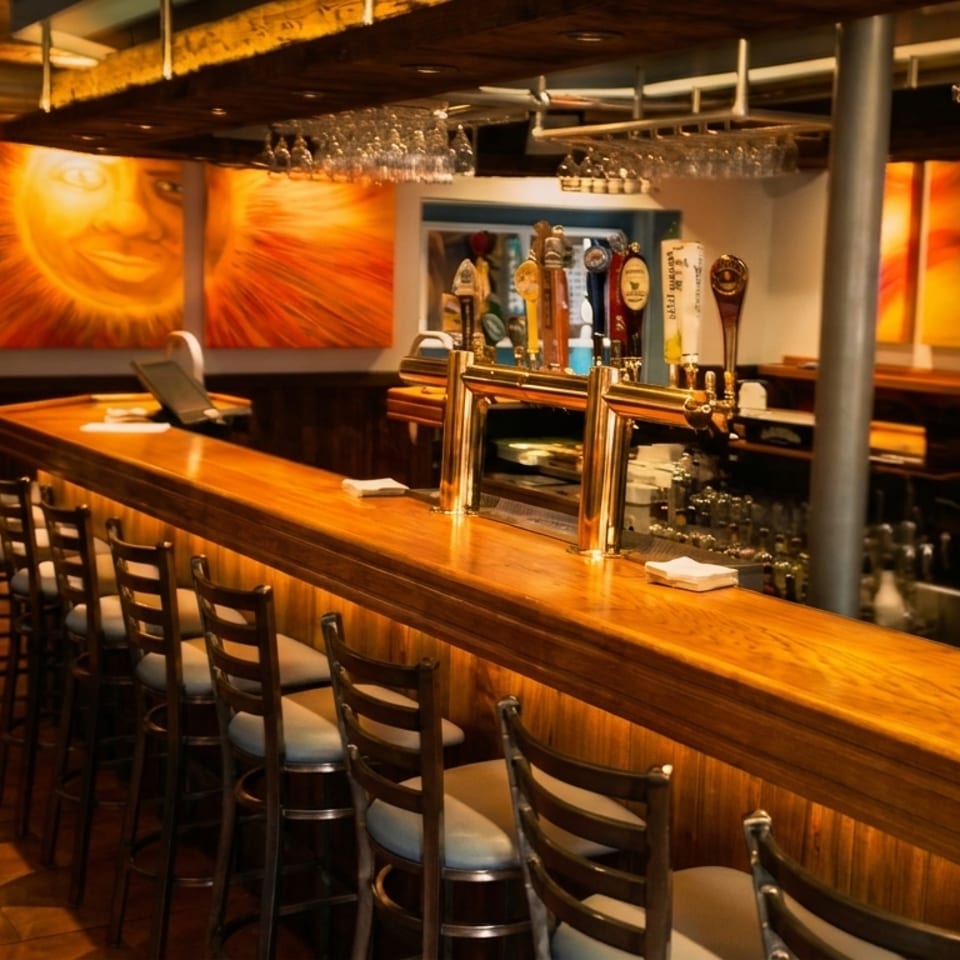Warmly lit restaurant bar with wooden counter, stools, beer taps, hanging glassware, colorful artwork, and television.
