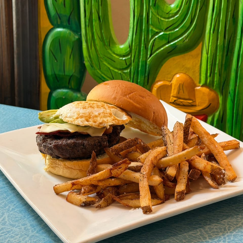 Cheeseburger with lettuce and tomato served alongside crispy fries on a white plate, against a colorful cactus mural.