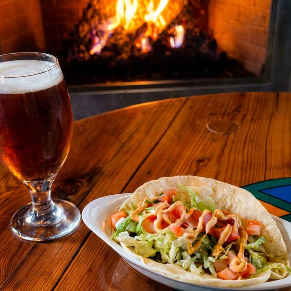 Taco filled with lettuce, tomatoes, and sauce beside a dark beer on a wooden table near a glowing fireplace.