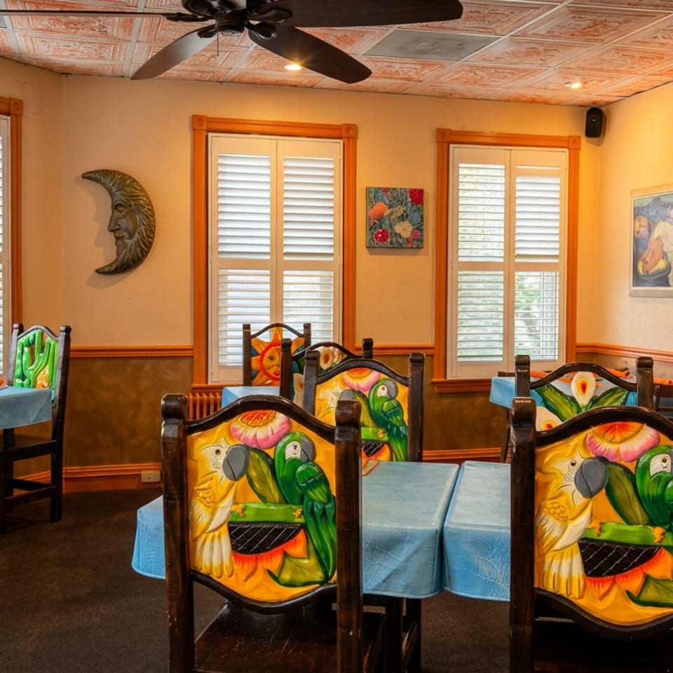 Warmly lit restaurant dining room with colorful parrot-painted chairs, blue tablecloths, shuttered windows, and decorative artwork.