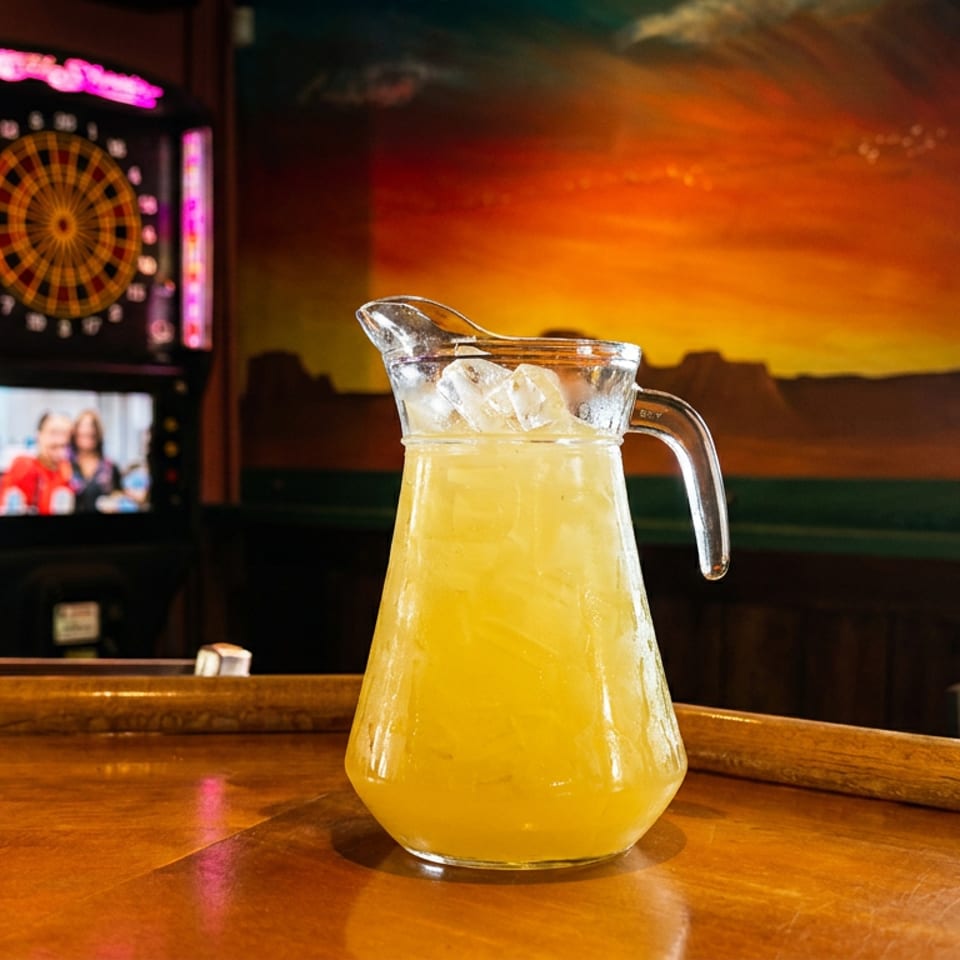 Glass pitcher of iced yellow beverage on a polished bar, with a dartboard and sunset mural in the restaurant background.