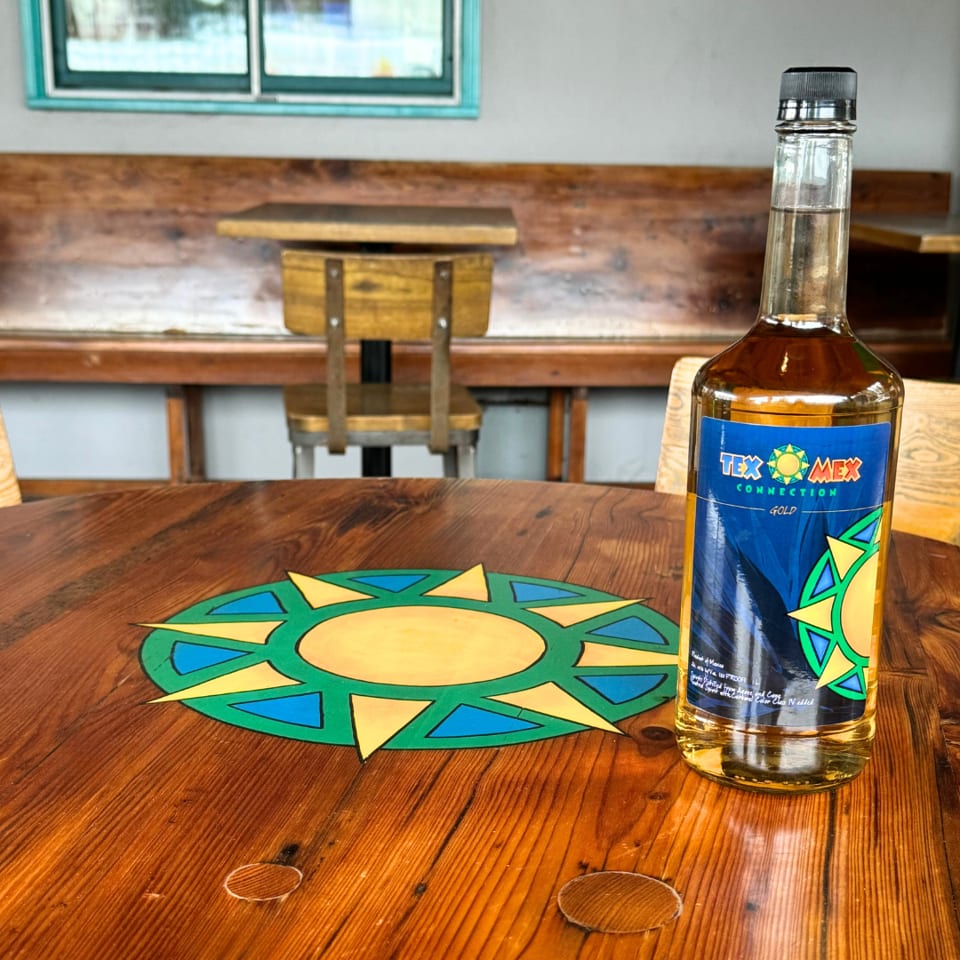 Bottle of Tex Mex Connection Gold liquor on a colorful sunburst placemat atop a rustic wooden restaurant table.