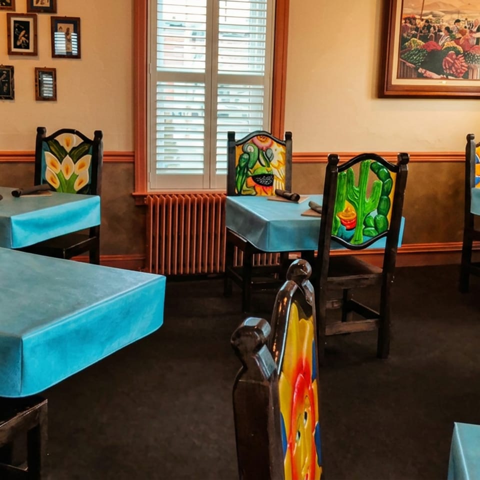 Colorful restaurant dining room with turquoise tablecloths, painted chairs, framed artwork, and shuttered windows.