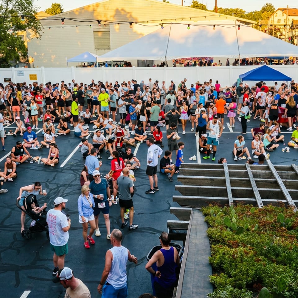 Crowded outdoor event on a rooftop parking deck, with string lights, tents, greenery, and surrounding urban buildings.