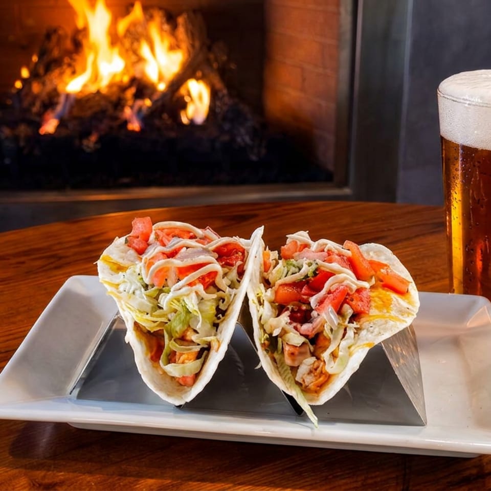 Two loaded tacos and a frothy pint of beer sit on a wooden table beside a glowing fireplace.