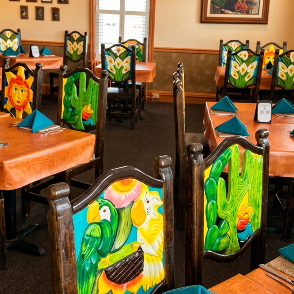Colorful restaurant dining room with tropical parrot-themed chairs, orange tablecloths, turquoise napkins, and framed artwork.